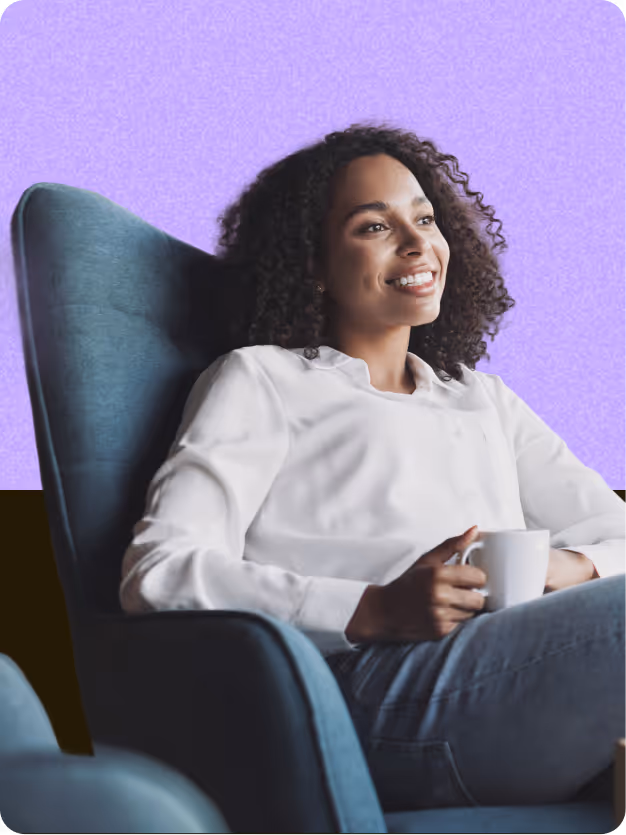 Smiling woman with curly hair holding a white mug seated in a blue chair against a purple background.