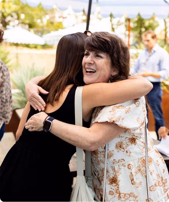 Two women warmly hugging outdoors, one wearing a floral dress and the other a black sleeveless top.