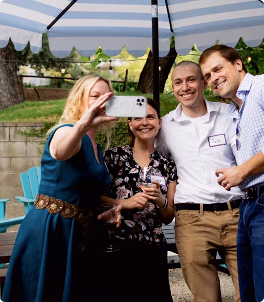 Four people smiling and posing for a selfie under a blue and white striped umbrella outdoors.