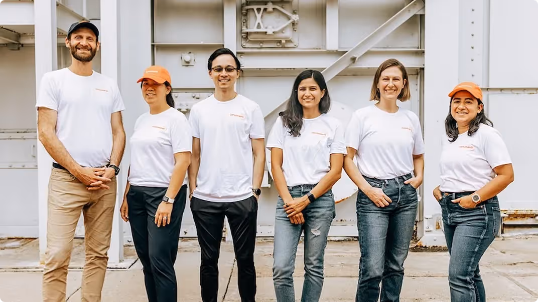Group of six smiling people standing outdoors wearing white t-shirts and casual pants.