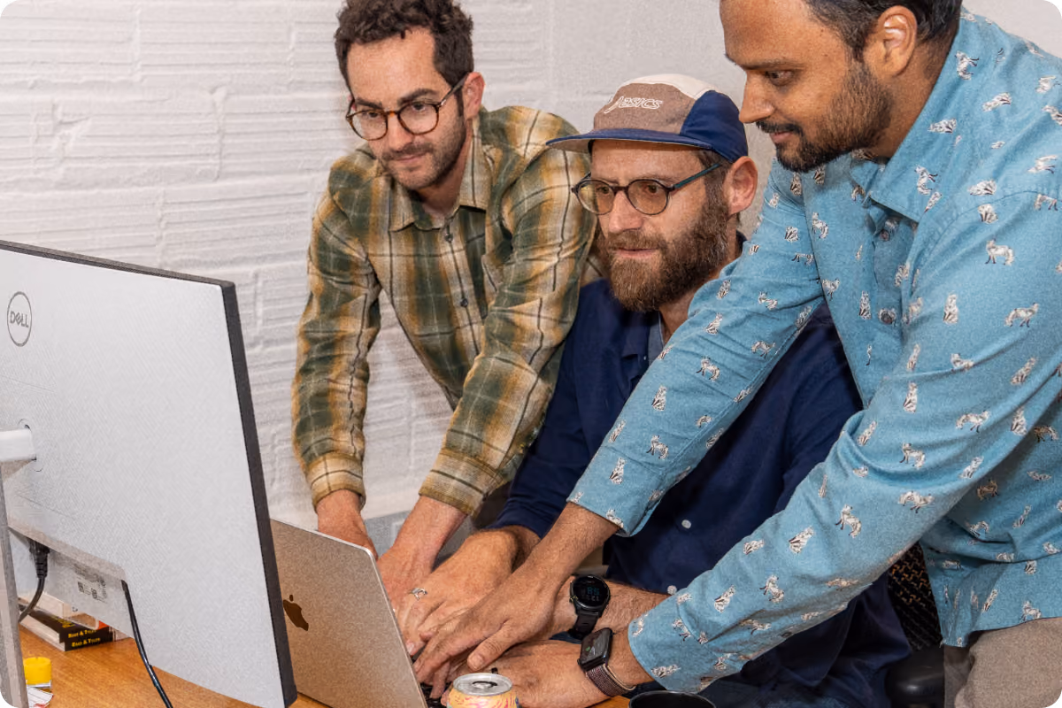 Three men closely collaborating while working on a laptop at a desk, with a large Dell monitor also on the desk.