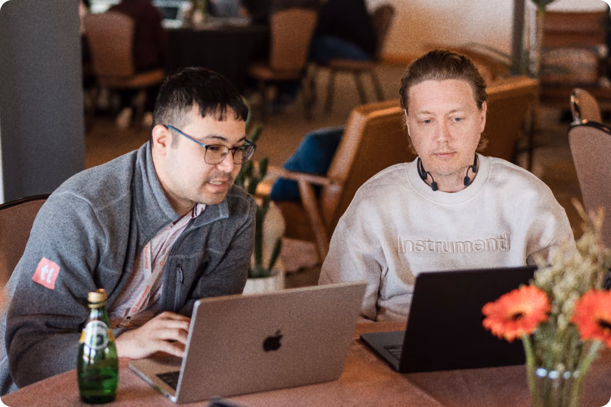 Two men sitting side by side at a table in a casual indoor setting, each working on a laptop. The man on the left wears glasses and a gray jacket with a red patch on the sleeve, focused on his silver MacBook. The man on the right wears a light-colored sweatshirt with the word 'Instrumentl' and headphones around his neck, looking at his dark laptop. A green glass bottle and a vase with orange flowers are on the table in the foreground.