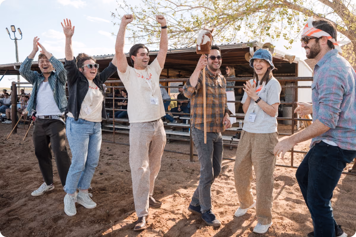 Six people outdoors on sandy ground celebrate excitedly, with some raising their arms and smiling. One person holds a stick with a horse head on top. They are casually dressed, with some wearing sunglasses and hats, standing near a metal fence and bleachers with other people in the background under a tree with sparse leaves.
