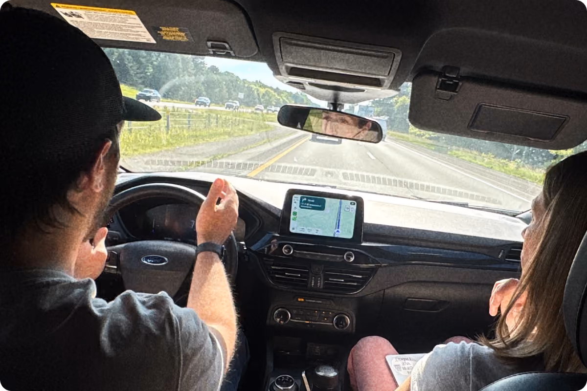 View from the back seat of a car showing a man driving and a woman sitting in the passenger seat, with a GPS navigation screen on the dashboard and a highway visible through the windshield.
