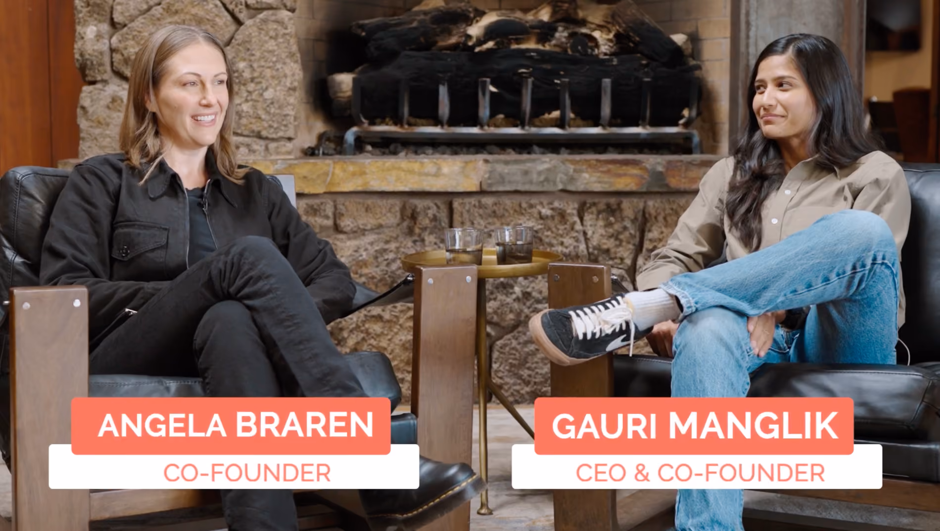 Two women seated on black leather chairs in front of a stone fireplace with a small round table between them holding two glasses. The woman on the left, Angela Braren, co-founder, wears a black jacket and black pants. The woman on the right, Gauri Manglik, CEO and co-founder, wears a light-colored shirt and blue jeans with crossed legs.