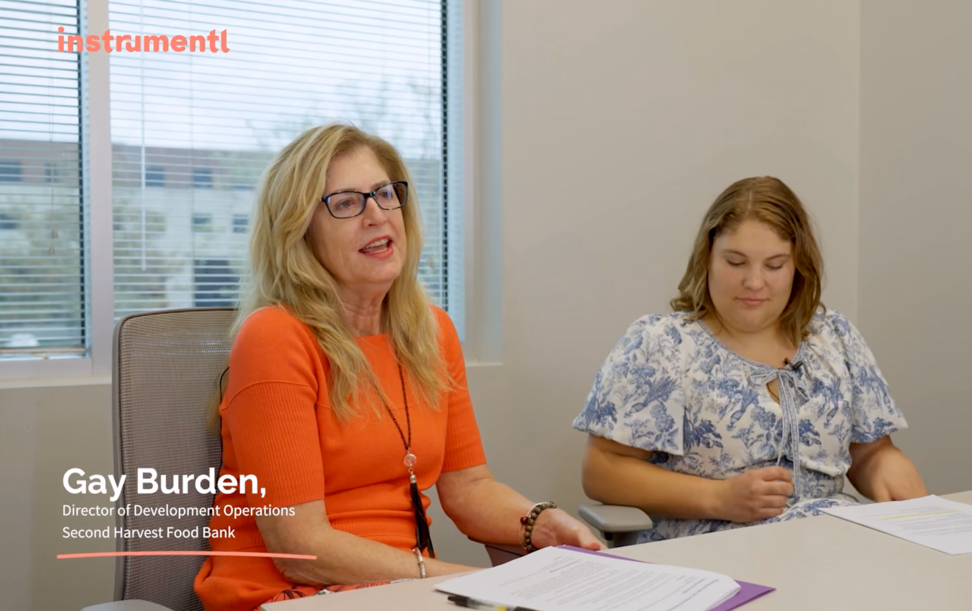 Two women seated at a table in an office room with window blinds; the woman on the left wears glasses and an orange top, the woman on the right wears a white floral blouse.