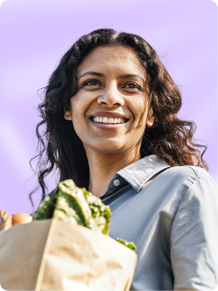 Smiling woman with curly dark hair holding a brown paper bag filled with fresh green lettuce and groceries, against a light green background with orange and purple decorative accents.