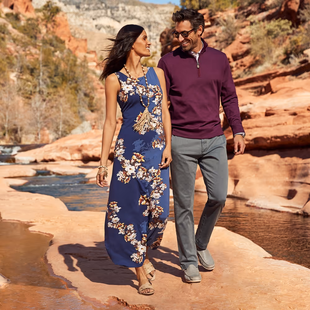 female model with tommy bahama dress and male model with jeans and maroon longsleeve walking in an orange rocky dessert by a stream 