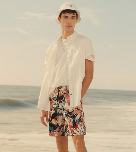 Young man on a beach wearing Tommy Hilfiger white shirt and bucket hat with tropical print shorts