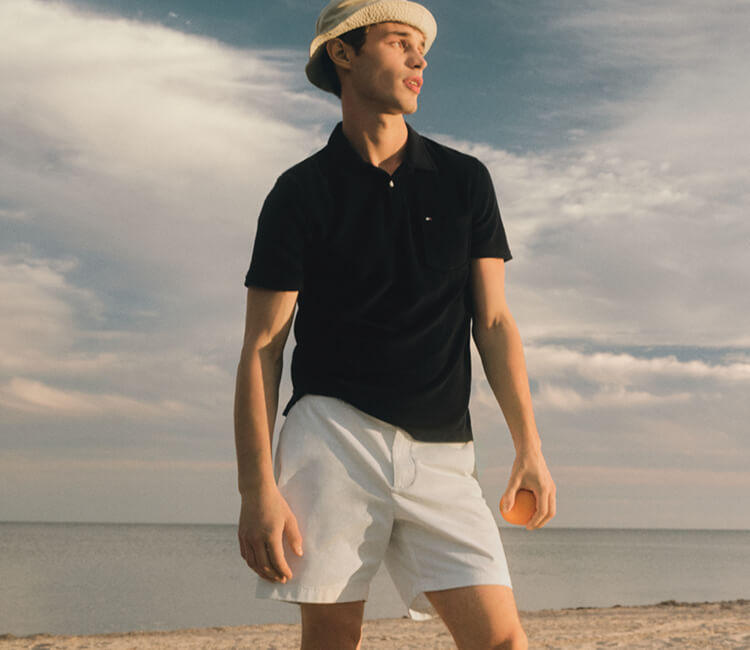Young man wearing Tommy Hilfiger Black collared short-sleeve shirt, white shorts, and tan bucket hat.