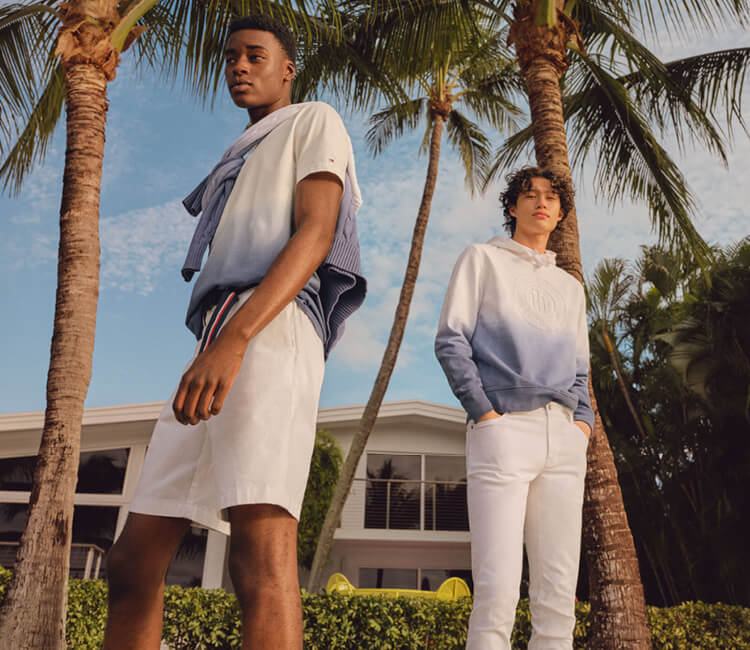 Two young men wearing Tommy Hilfiger blue and white gradient sweaters; one with white shorts and the other with white pants
