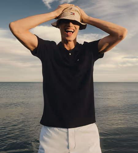 Young man wearing Tommy Hilfiger tan bucket hat, black collared short-sleeved shirt, and white shorts