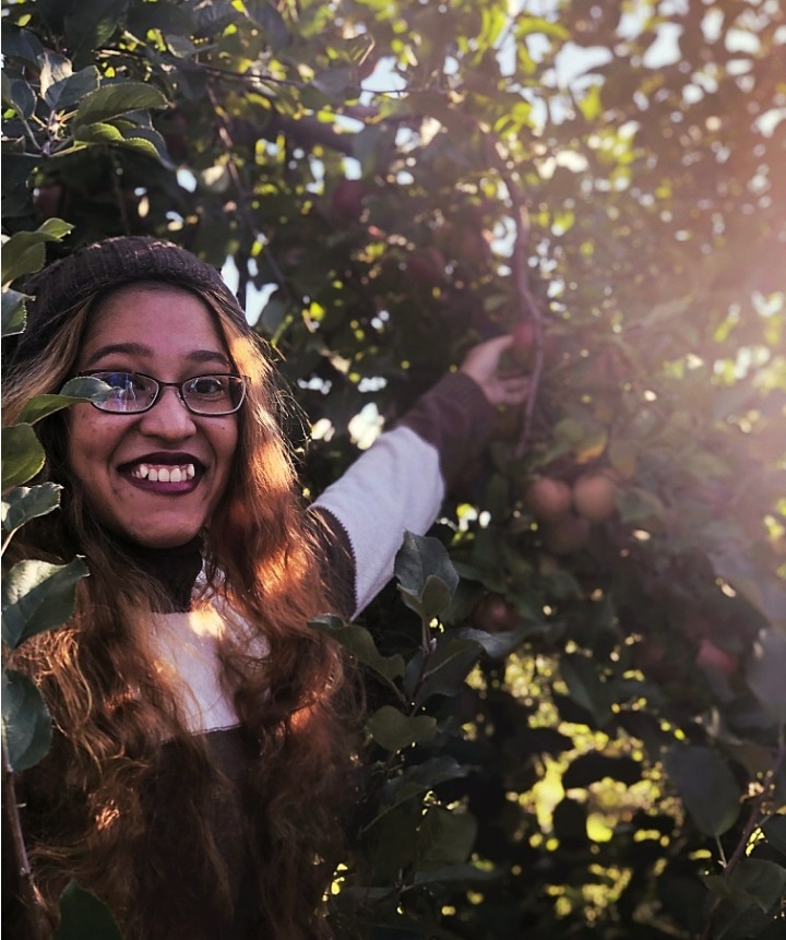A picture of me smiling and picking an apple in a brown sweater and beanie