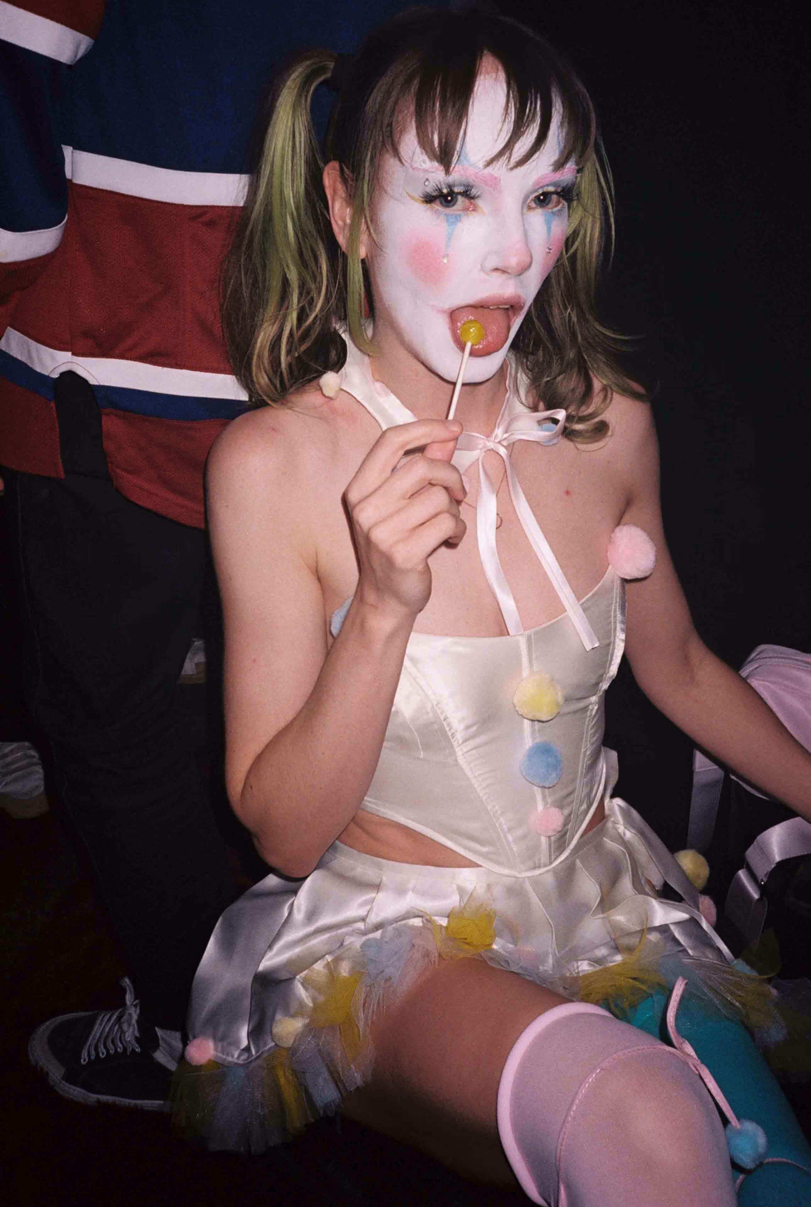 A young girl dressed as a clown eating a lollipop.
