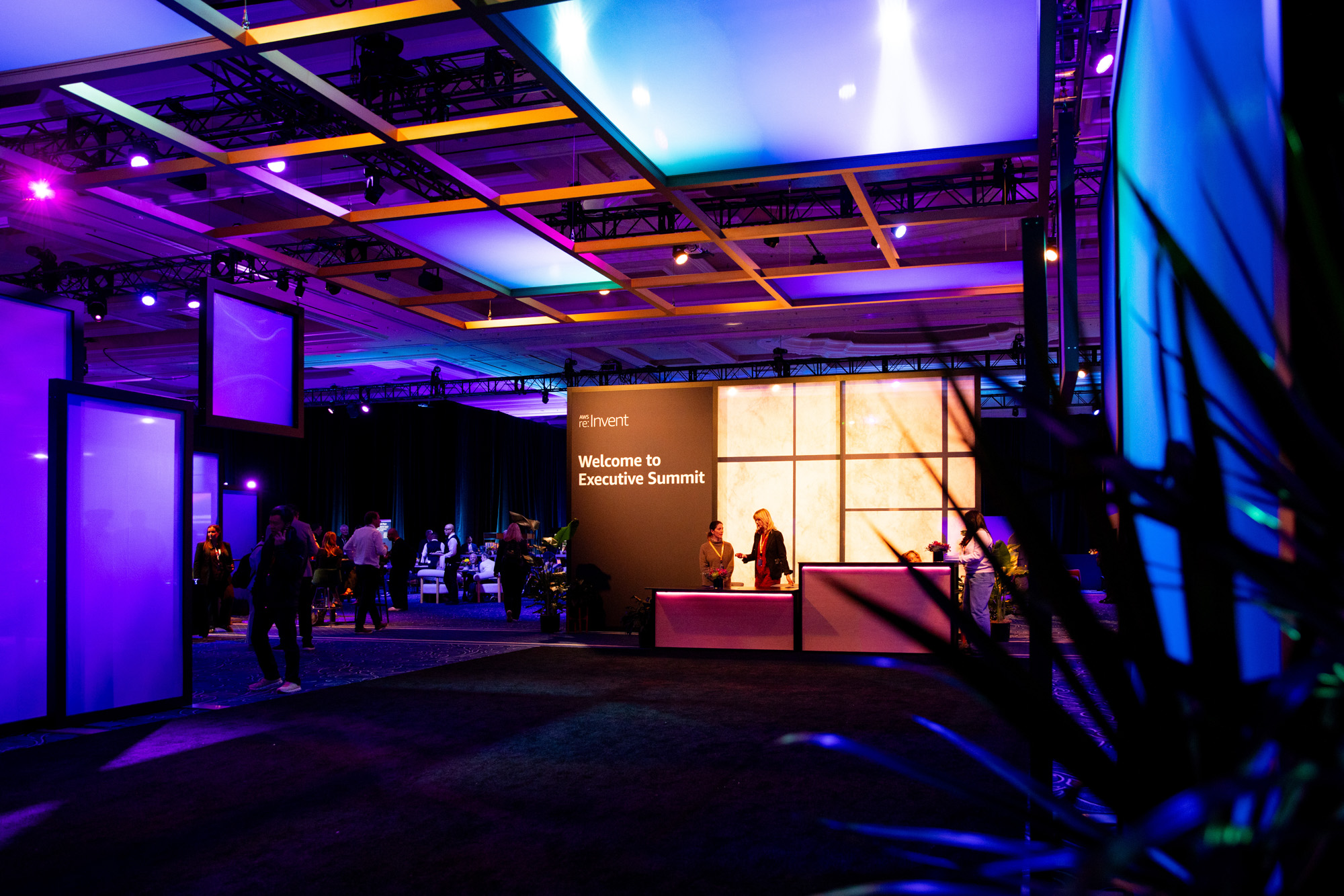 Dimly lit room with people gathered around tables and a welcome desk displaying 'Welcome to Executive Summit' at an AWS re:Invent event.
