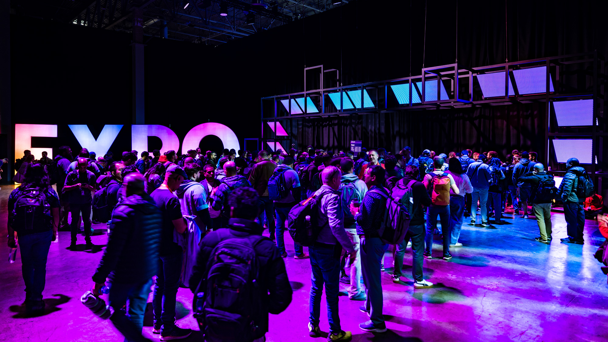 Crowd of people waiting in line inside a dimly lit expo hall with colorful purple and blue lighting.