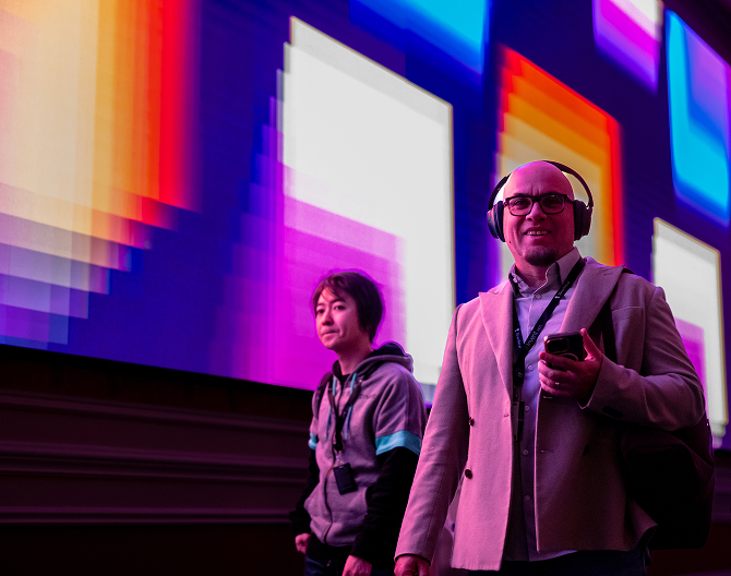 Two men walking indoors in front of a large colorful digital screen, one wearing headphones and holding a phone.