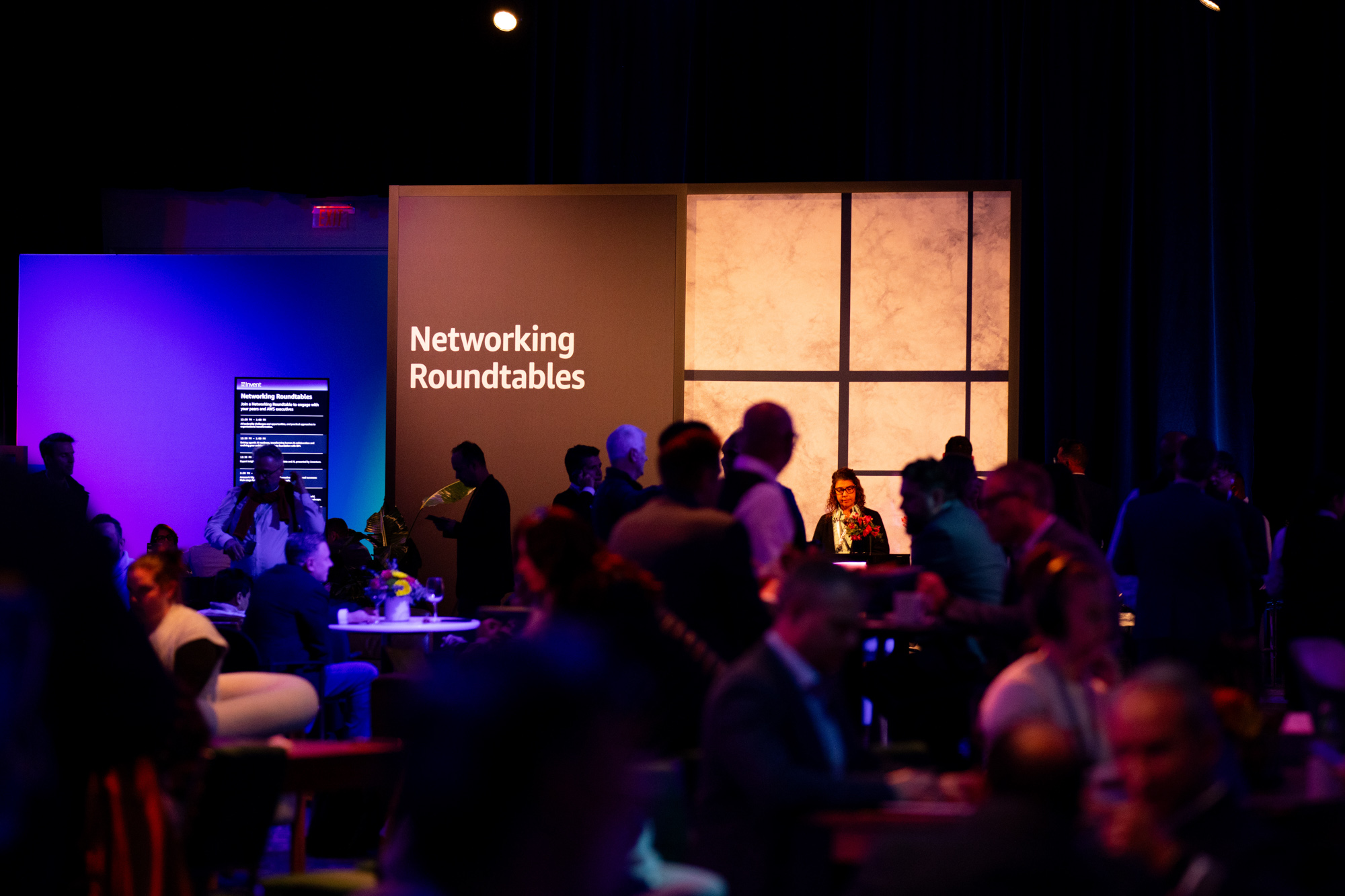 People gathered in a dimly lit room for networking roundtables with a sign and a woman at a reception desk.