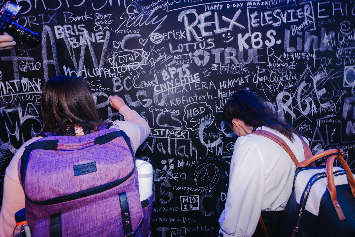 Two people with backpacks writing on a blackboard wall covered with various white chalk signatures and messages.