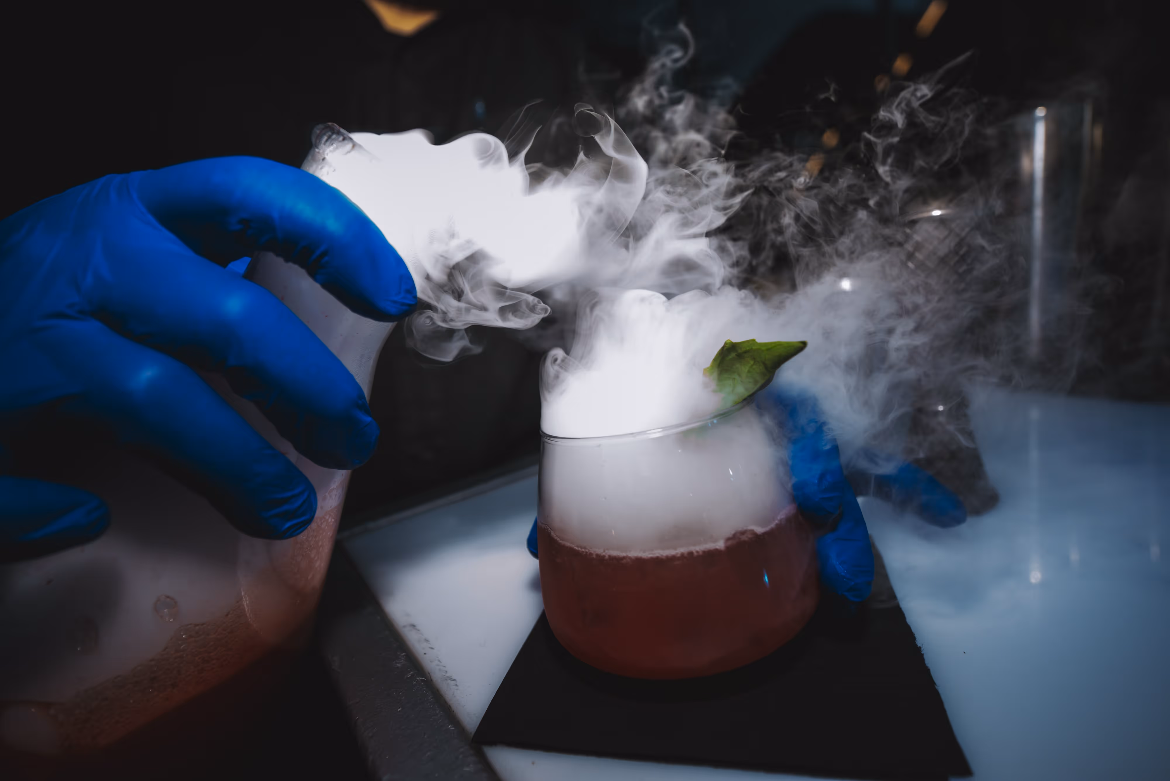 A person in blue gloves holding a glass with smoke coming out of it.