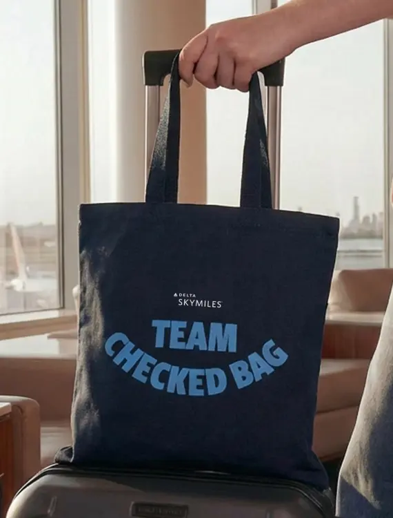 Right image of the blue Delta ‘Team Checked Bag’ tote bag, resting on a suitcase in the SkyClub lounge.