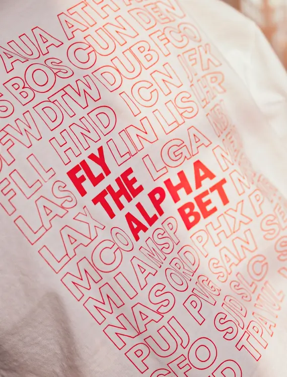 Middle image of Delta’s ‘Fly The Alphabet” crewneck white t-shirt with red text.