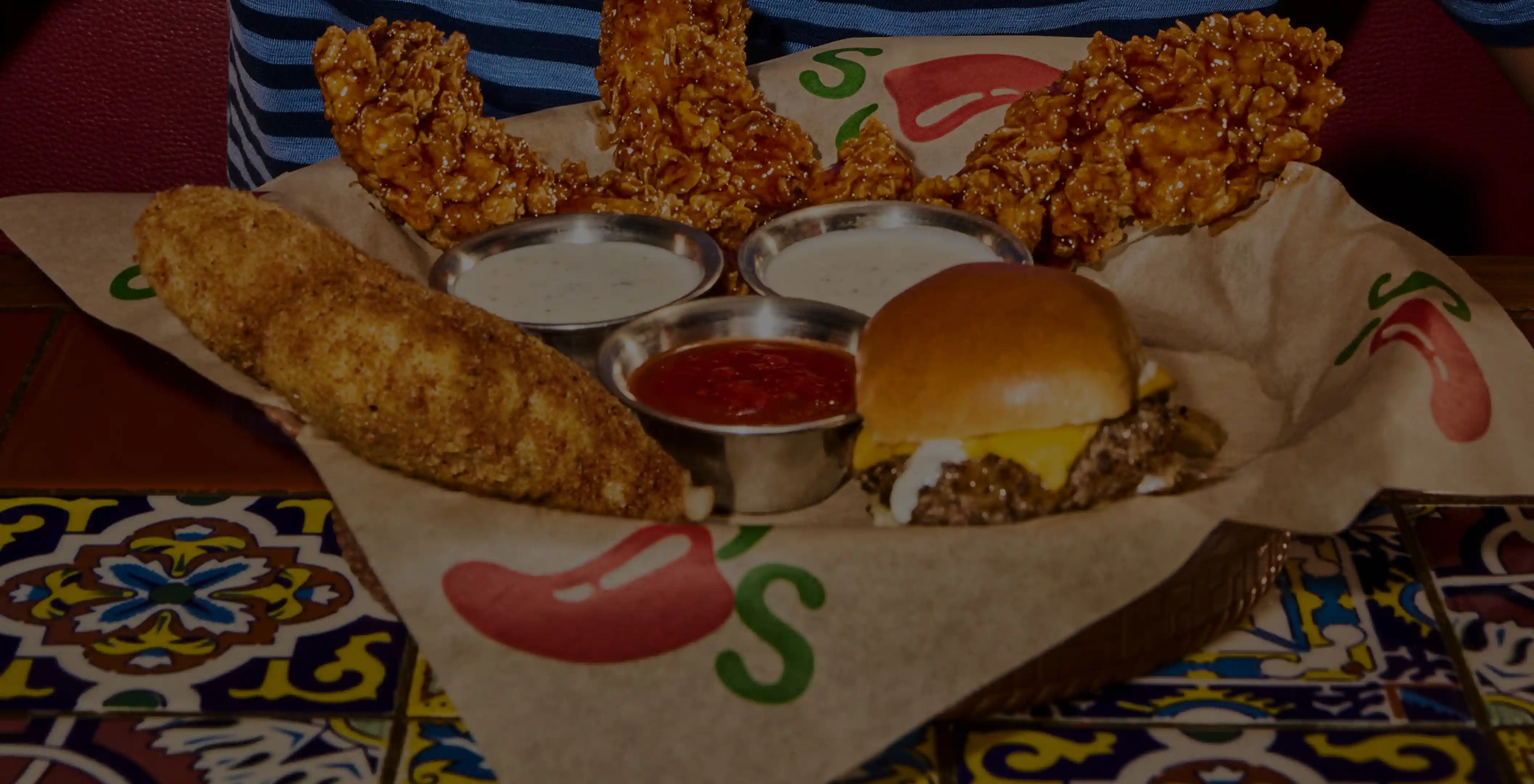 Basket of Chili’s chicken tenders, corn, dipping sauces, and cheeseburger on a colorful table.
