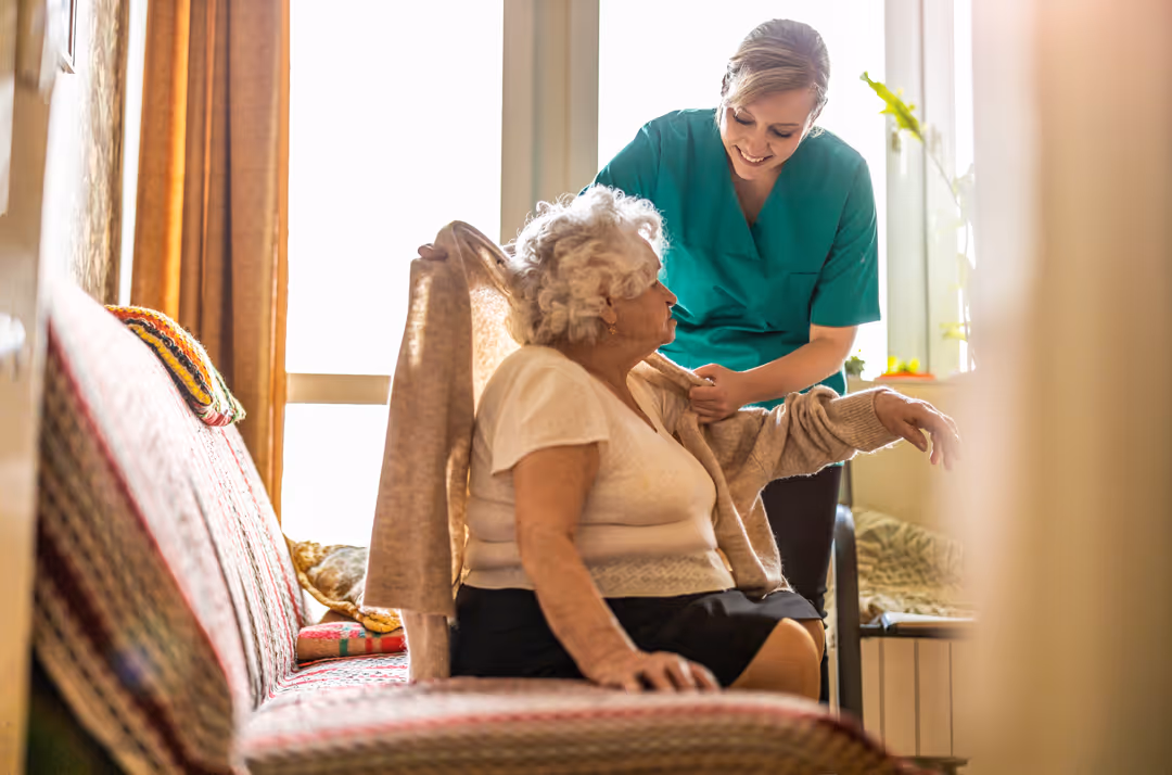 A female nurse helping an elderly patient put on a sweater inside of her home.