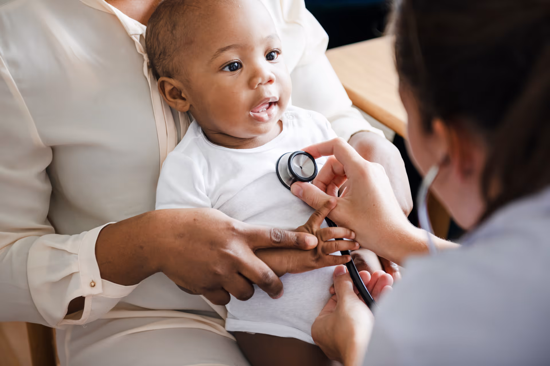 A doctor checking the heartbeat of a baby. The baby is being held in their mothers arms while the doctor closely listens in with her stethoscope.