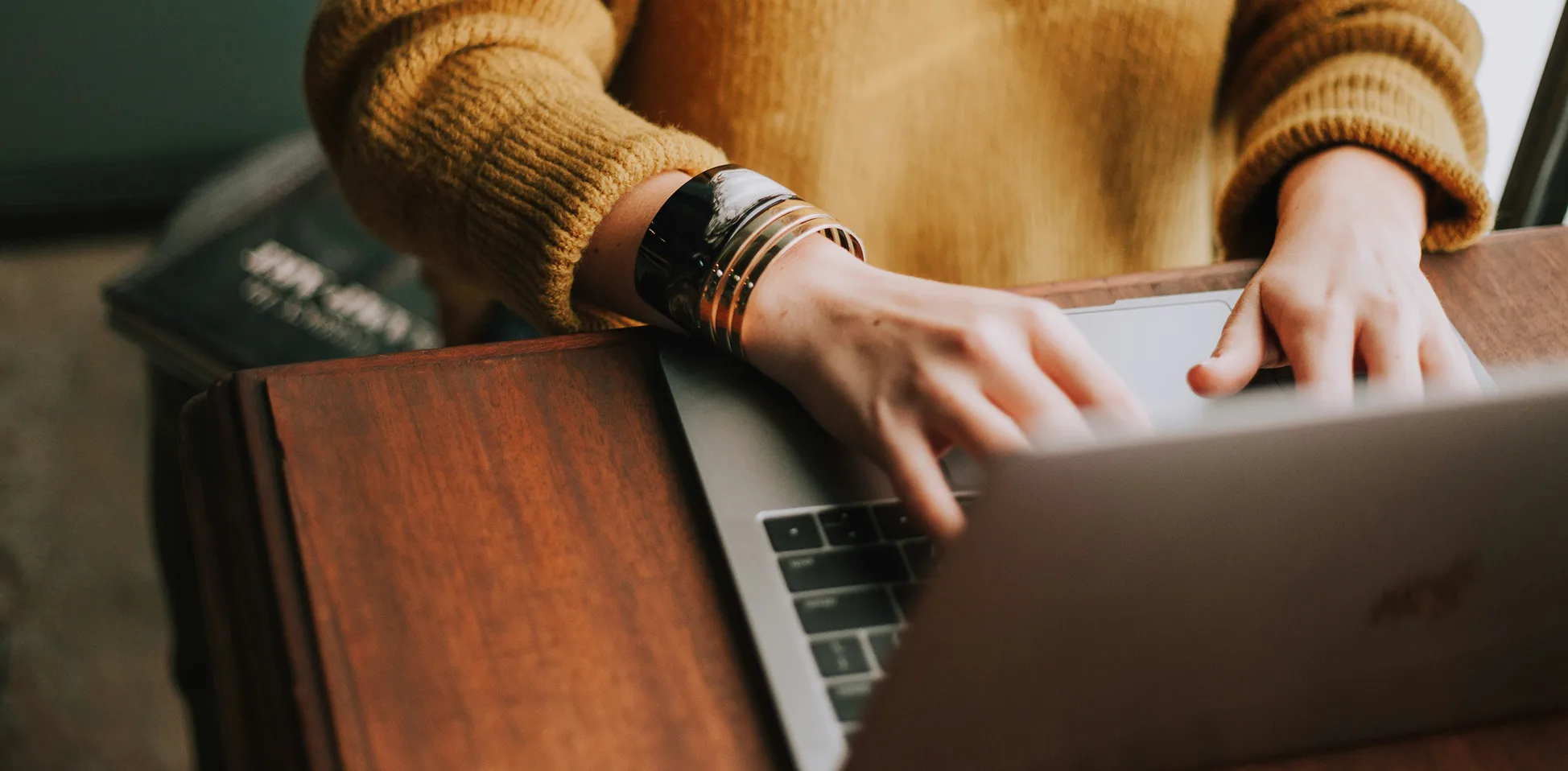 Person wearing a mustard sweater typing on a laptop at a wooden desk, with bracelets visible on their wrist.