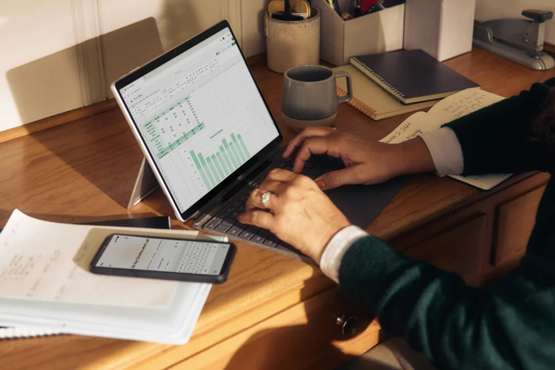 A person works on a spreadsheet at a wooden desk, with a phone, notebook, and coffee mug nearby.