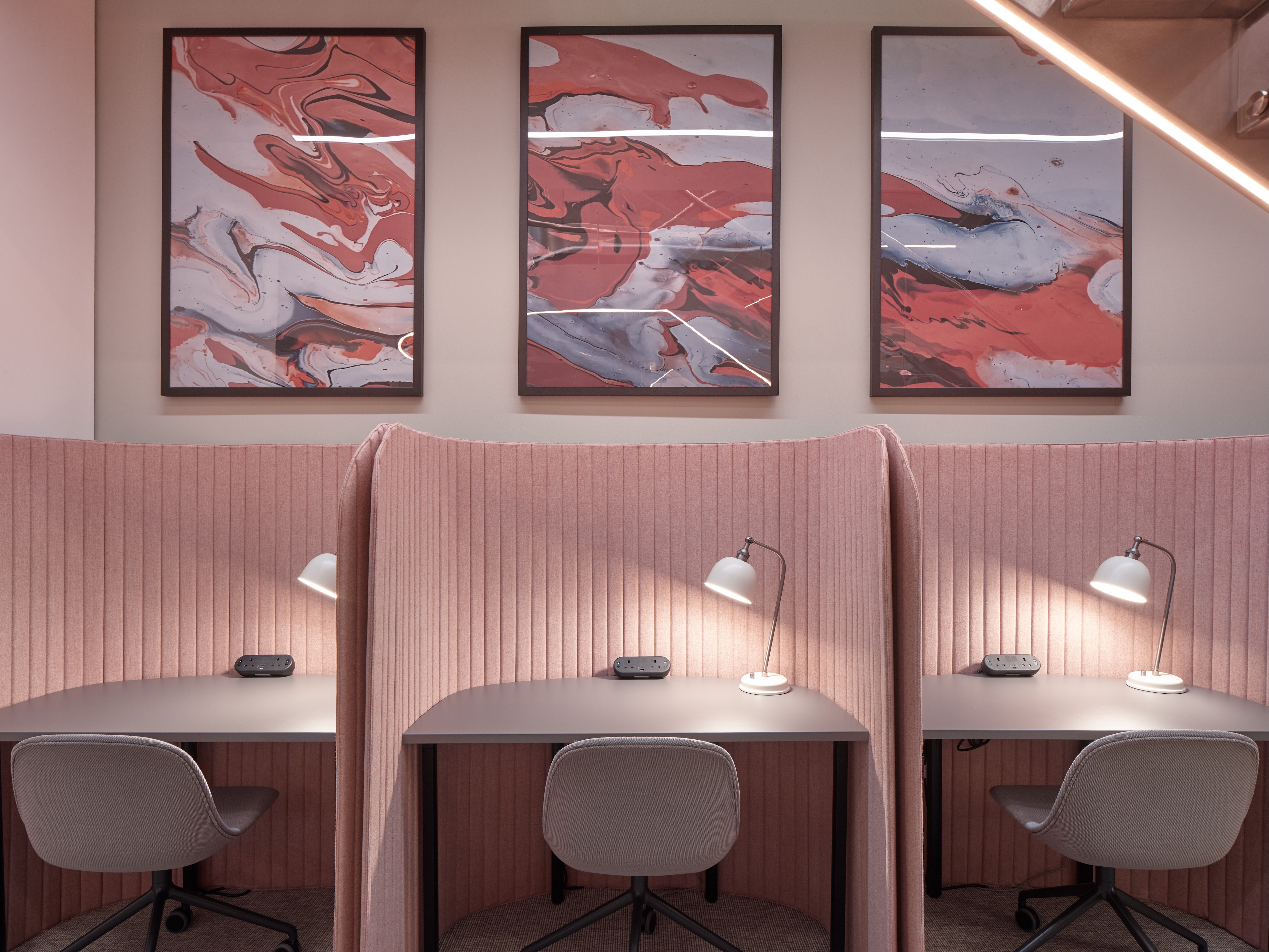 Three pink semi-private workstations with grey chairs, white desk lamps, and abstract red and grey artwork above.