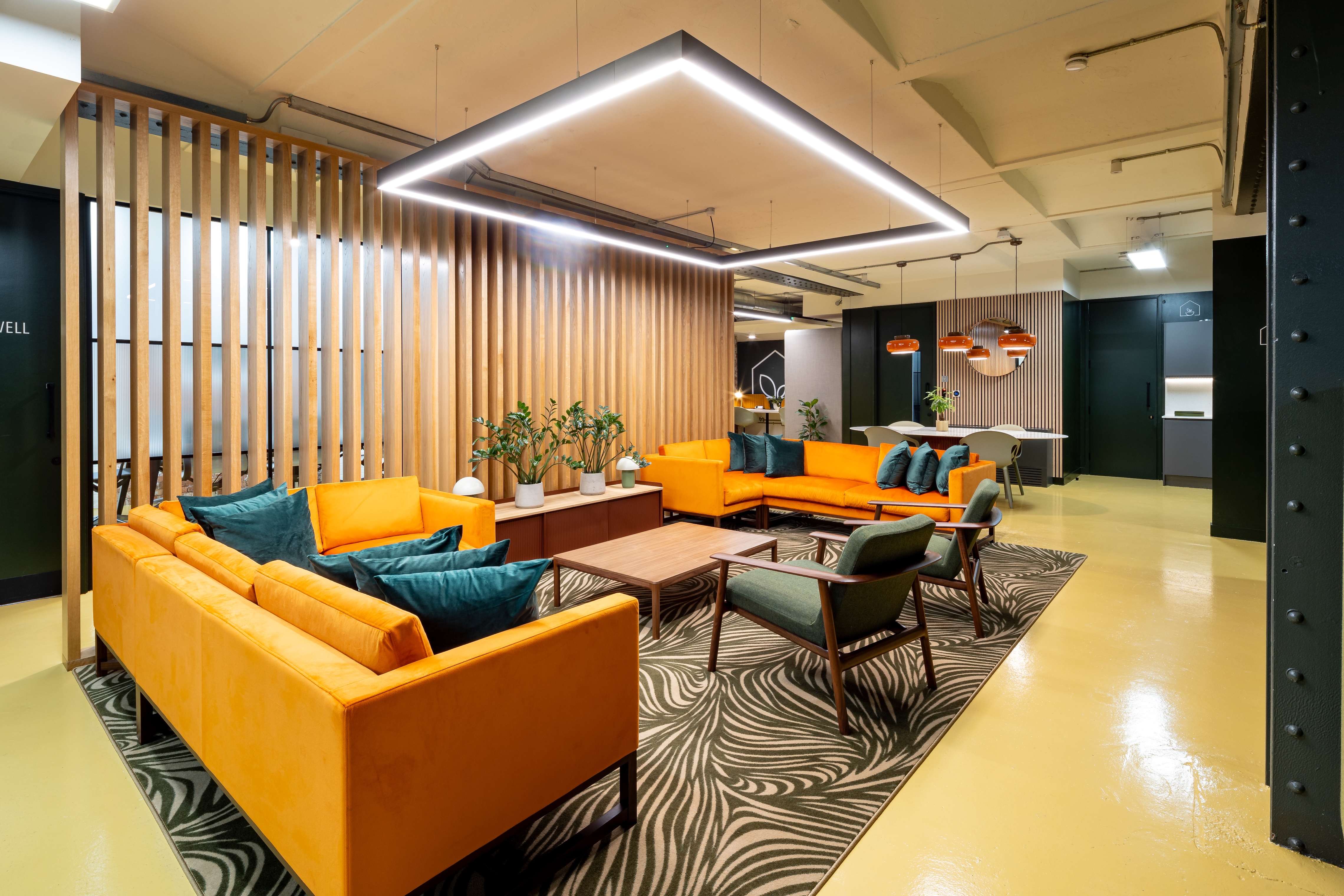 Modern office lounge with bright orange sofas, green armchairs, a patterned rug, wooden slat divider, and a square ceiling light fixture.