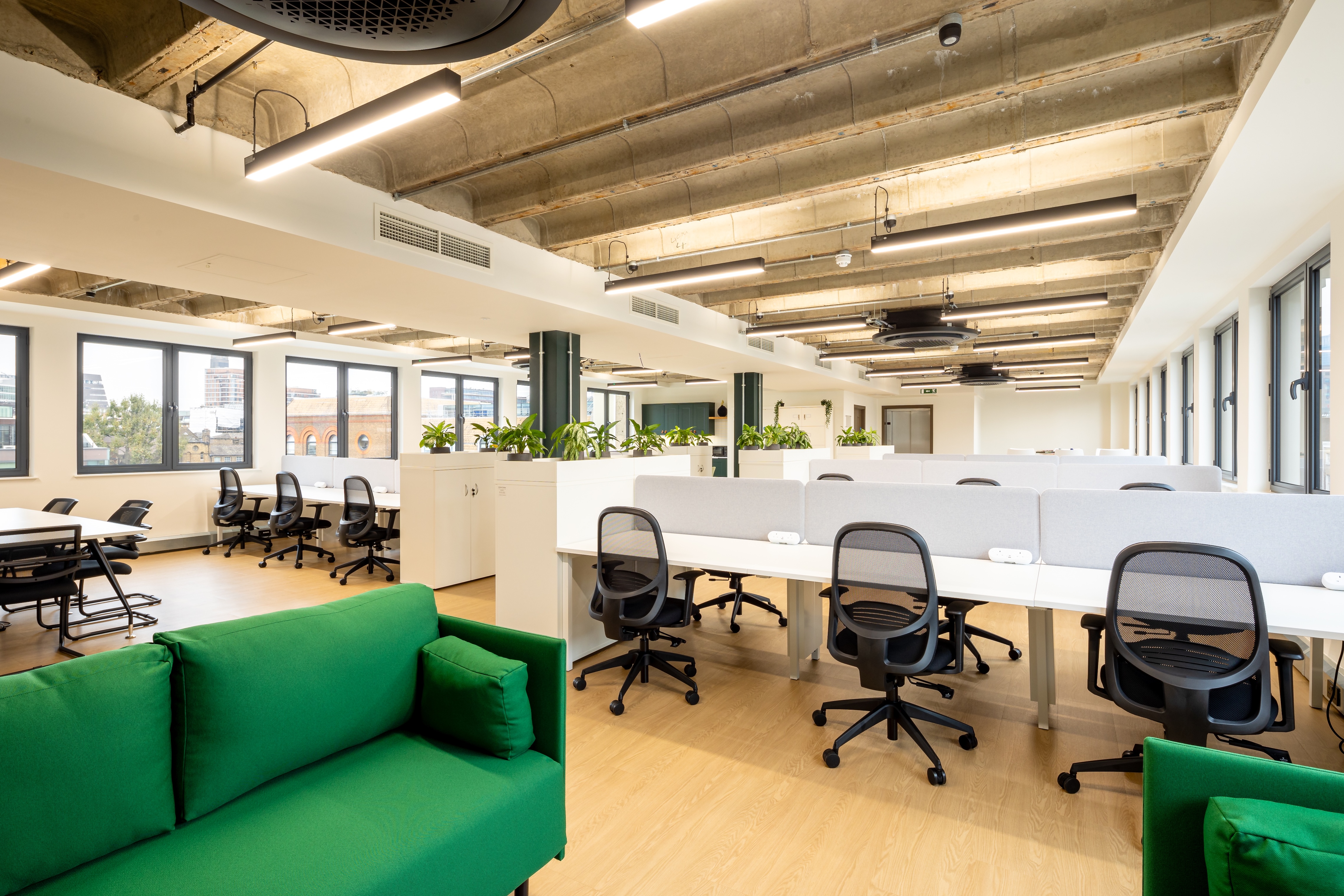 Modern open-plan office with rows of white desks, black mesh office chairs, and green sofas in the foreground under exposed concrete ceiling with hanging lights.