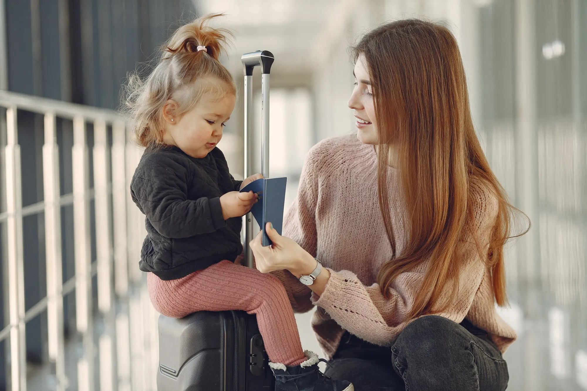 Mother and toddler at the airport, with the child sitting on a suitcase and holding a passport as the mother looks on smiling