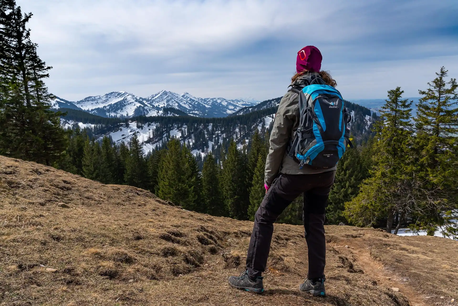 A woman with braided hair is seen from behind, hiking through a lush green valley surrounded by tall mountains. She carries a large yellow backpack with a baseball cap attached and walks along a trail dotted with wildflowers under a cloudy sky.