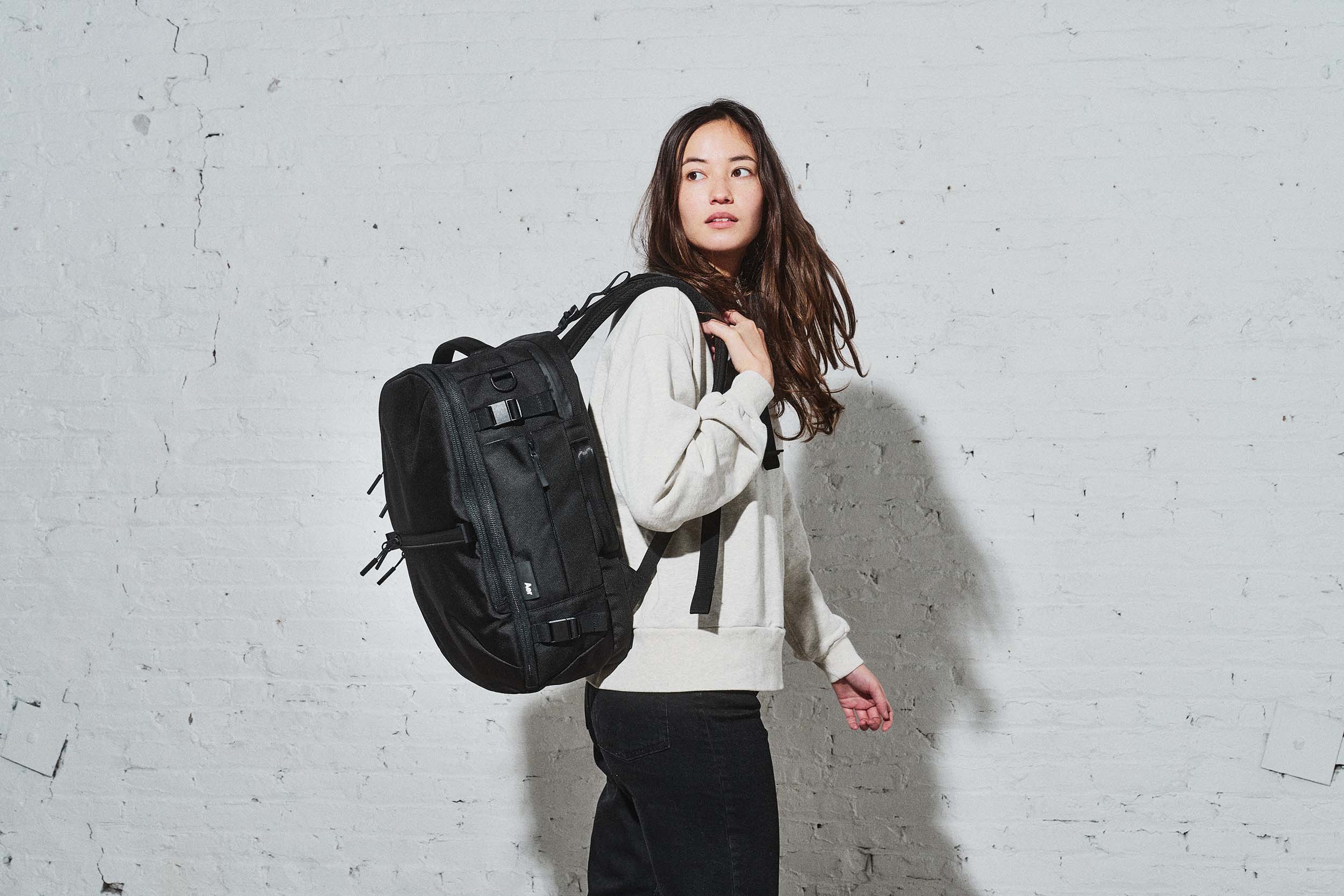 A woman wearing a light grey sweatshirt and black pants stands in front of a white brick wall, carrying a black Aer Travel Pack 3 Small backpack over one shoulder. She looks off to the side with a neutral expression in a well-lit, minimalist indoor setting.
