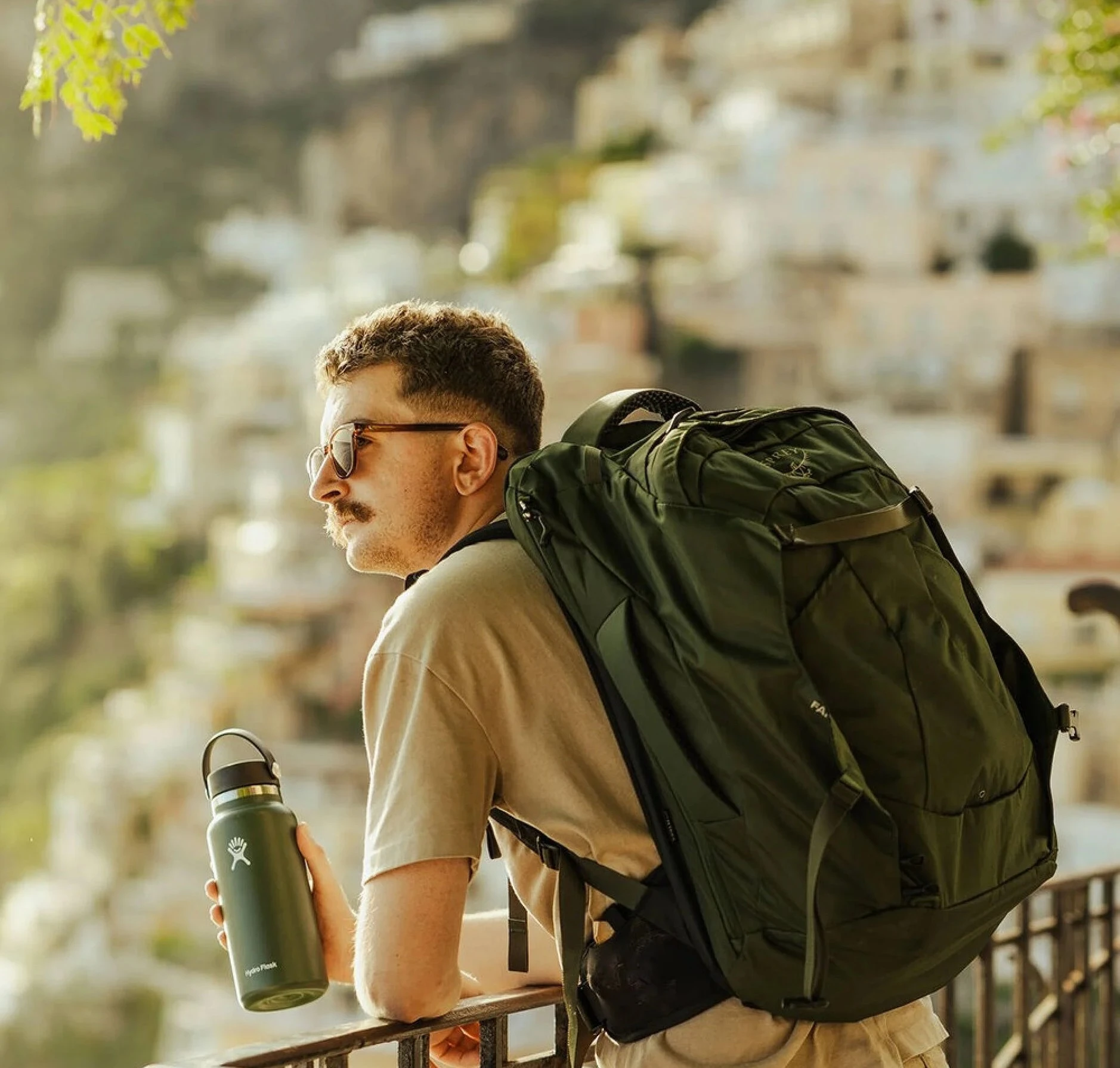 A man with a mustache wearing sunglasses and a tan t-shirt leans on a railing overlooking a scenic hillside town. He carries a dark green Osprey Farpoint 40L backpack and holds a green Hydro Flask water bottle. The background features sunlit, blurred buildings on a hillside, suggesting a Mediterranean or coastal setting.