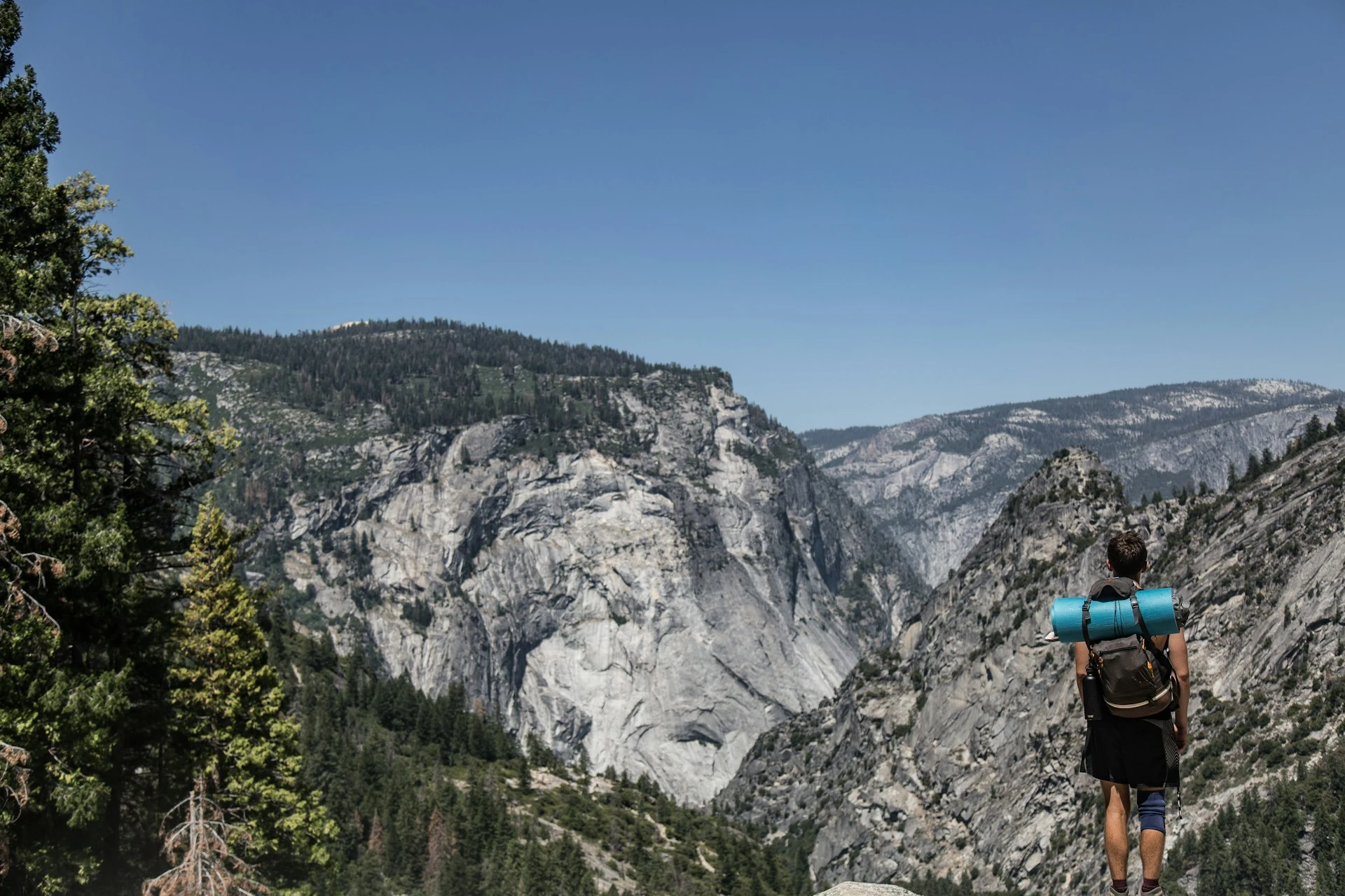A hiker with a blue sleeping mat strapped to their backpack stands on a rocky ledge, overlooking a rugged mountain landscape under a clear blue sky. The scene showcases steep granite cliffs, dense green forests, and distant mountain ranges.