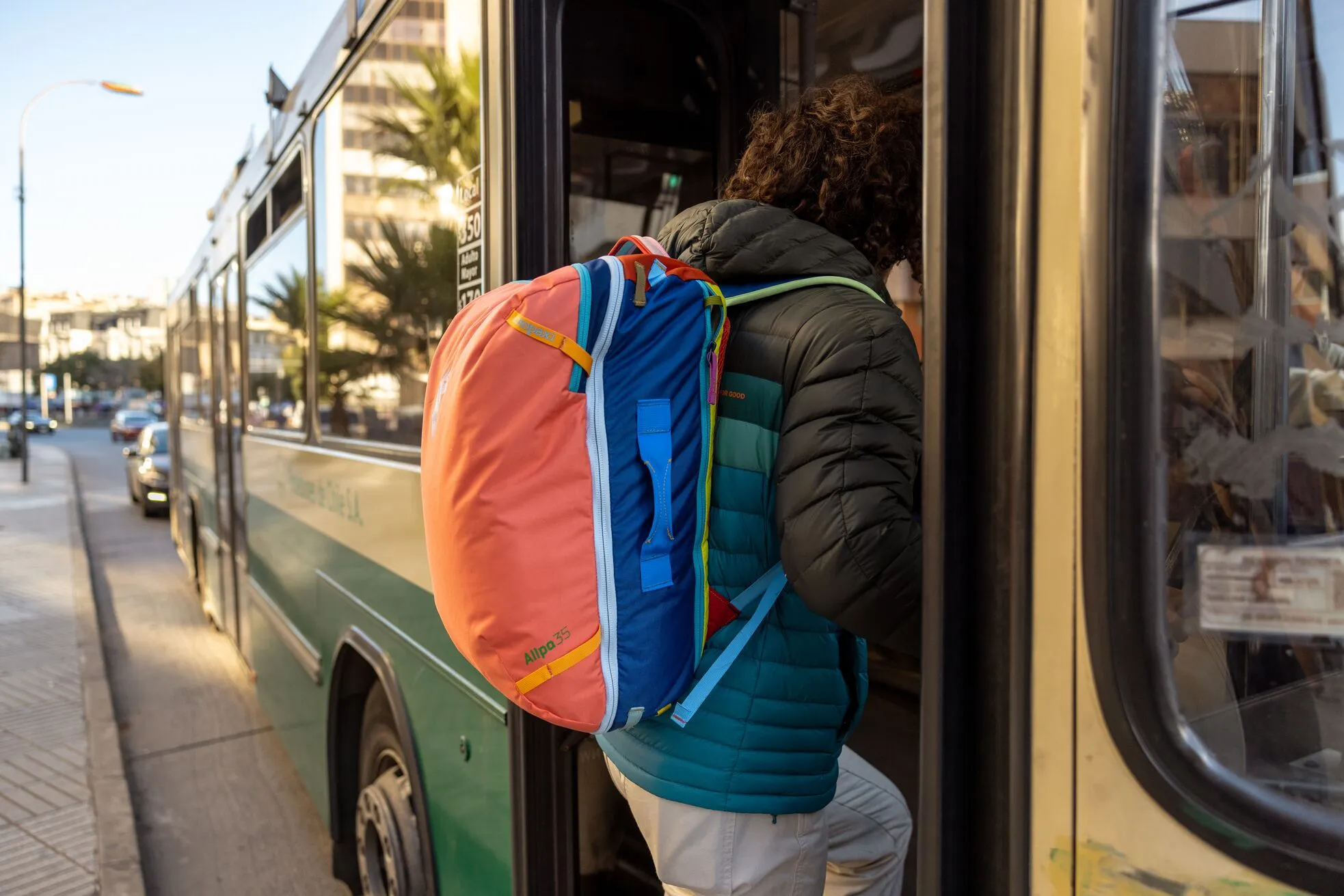 A person wearing a teal and black puffer jacket is boarding a city bus while carrying a colorful Cotopaxi Allpa 35L backpack in coral, blue, and yellow. The urban setting includes cars, buildings, and palm trees visible in the background.