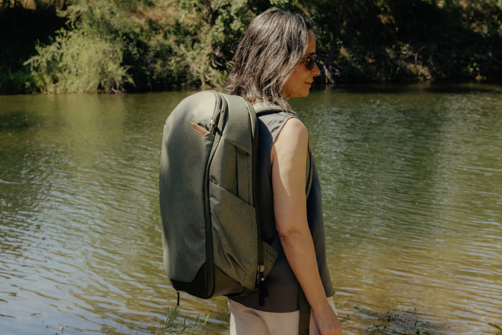 A person wearing sunglasses and a sleeveless top stands beside a calm river, carrying an olive green Peak Design Travel Backpack 30L. The natural outdoor setting features water, grass, and trees in the background, evoking a peaceful, adventure-ready vibe.
