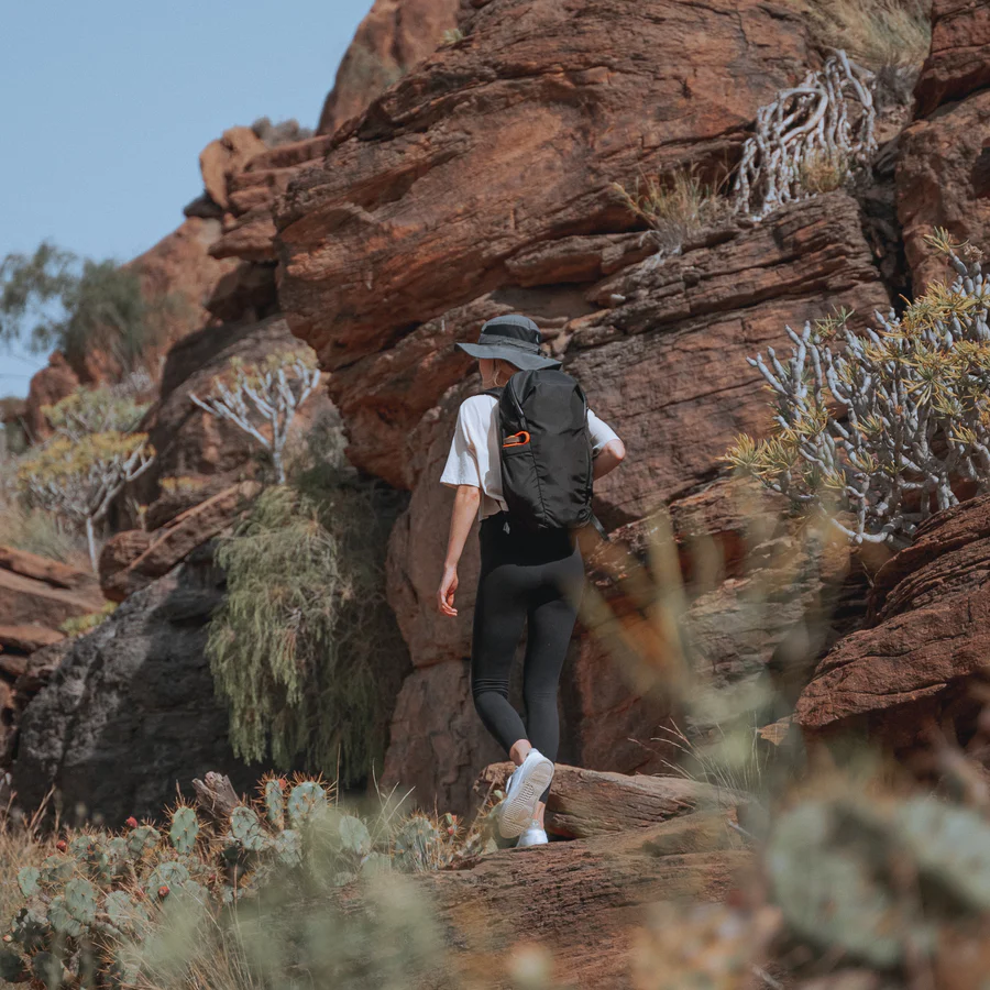 A person wearing a sun hat, white shirt, and black leggings hikes through a rocky desert landscape, carrying a black Able Carry Daybreaker 2 backpack. The environment features red rock formations, cacti, and desert shrubs under a clear blue sky.