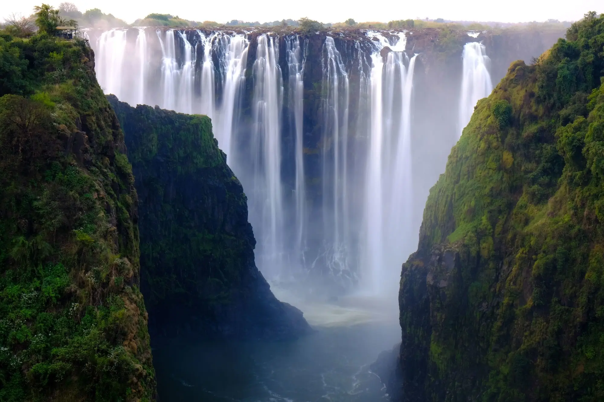 Victoria Falls cascading between lush, green cliffs, with mist rising from the powerful waterfall and trees lining the top edge