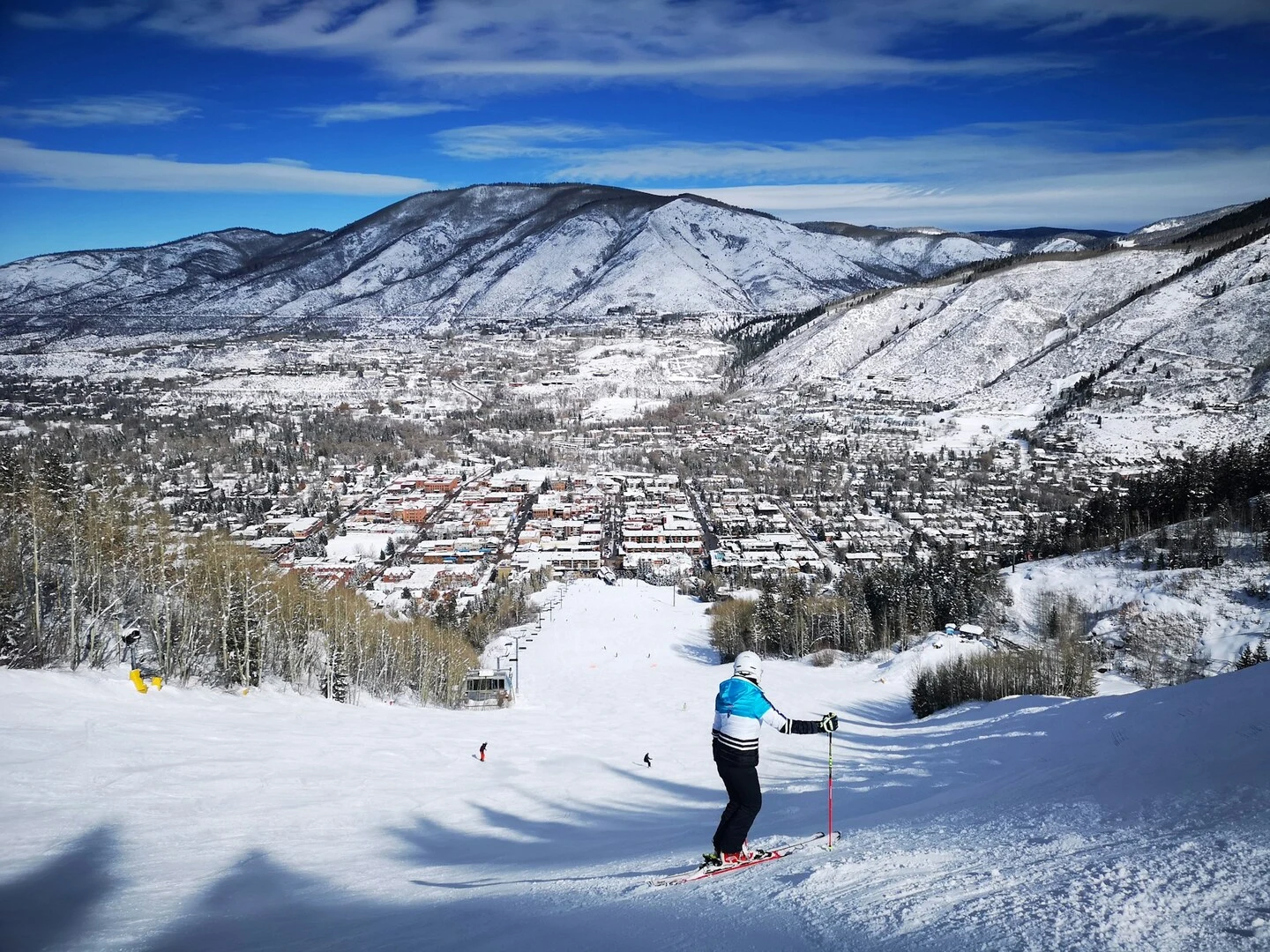 Skier descending a wide groomed run at Aspen Snowmass, Colorado, with a snowy mountain town and surrounding alpine peaks under a clear blue winter sky.