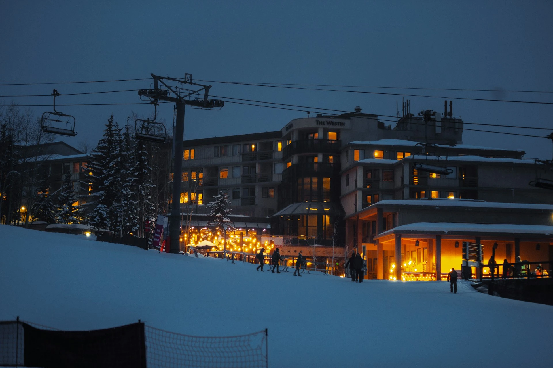 A group of people walk across a snow-covered path beside a softly lit building in Snowmass Colorado
