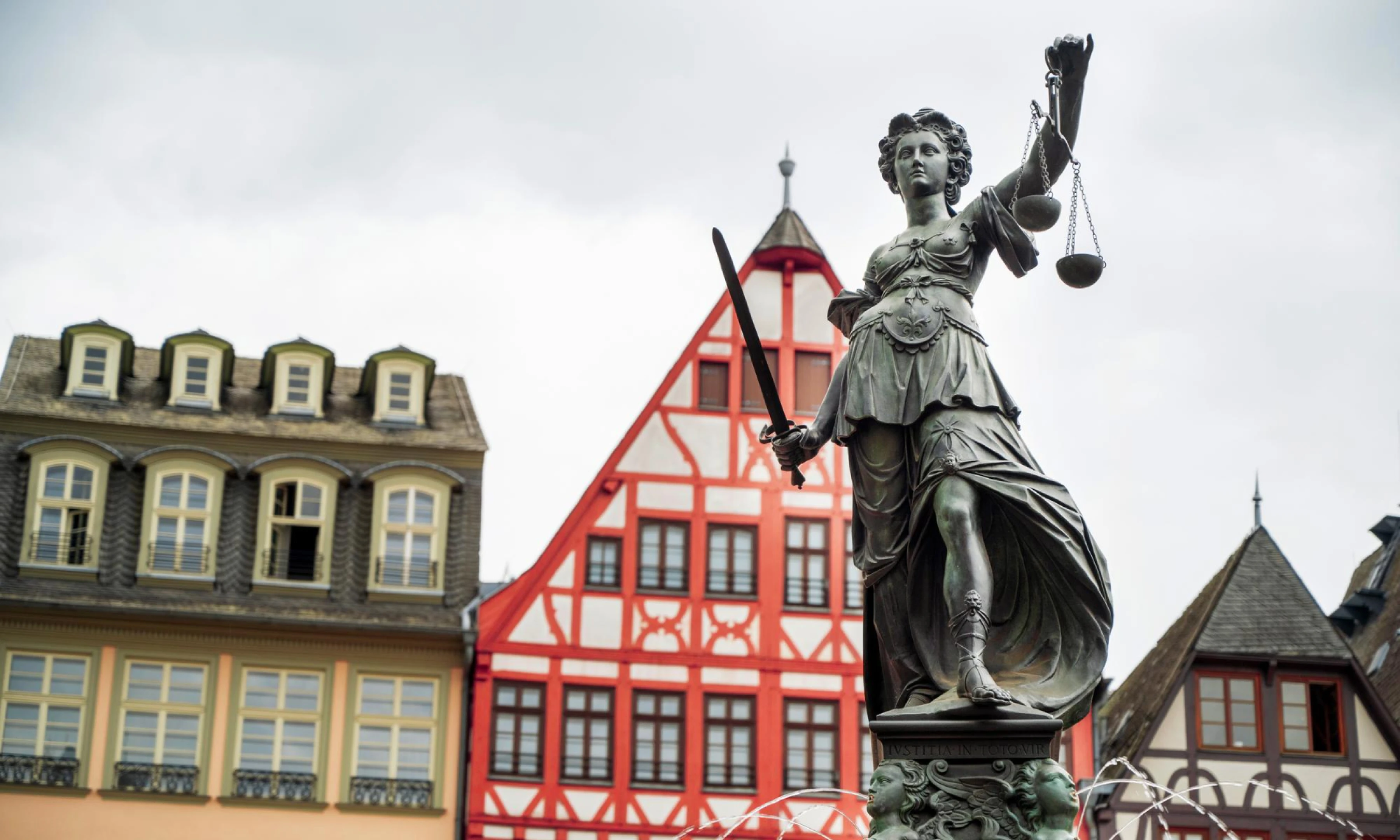 Statue of Lady Justice holding a sword and scales, located in Frankfurt's Römer Square, with traditional half-timbered buildings in the background.
