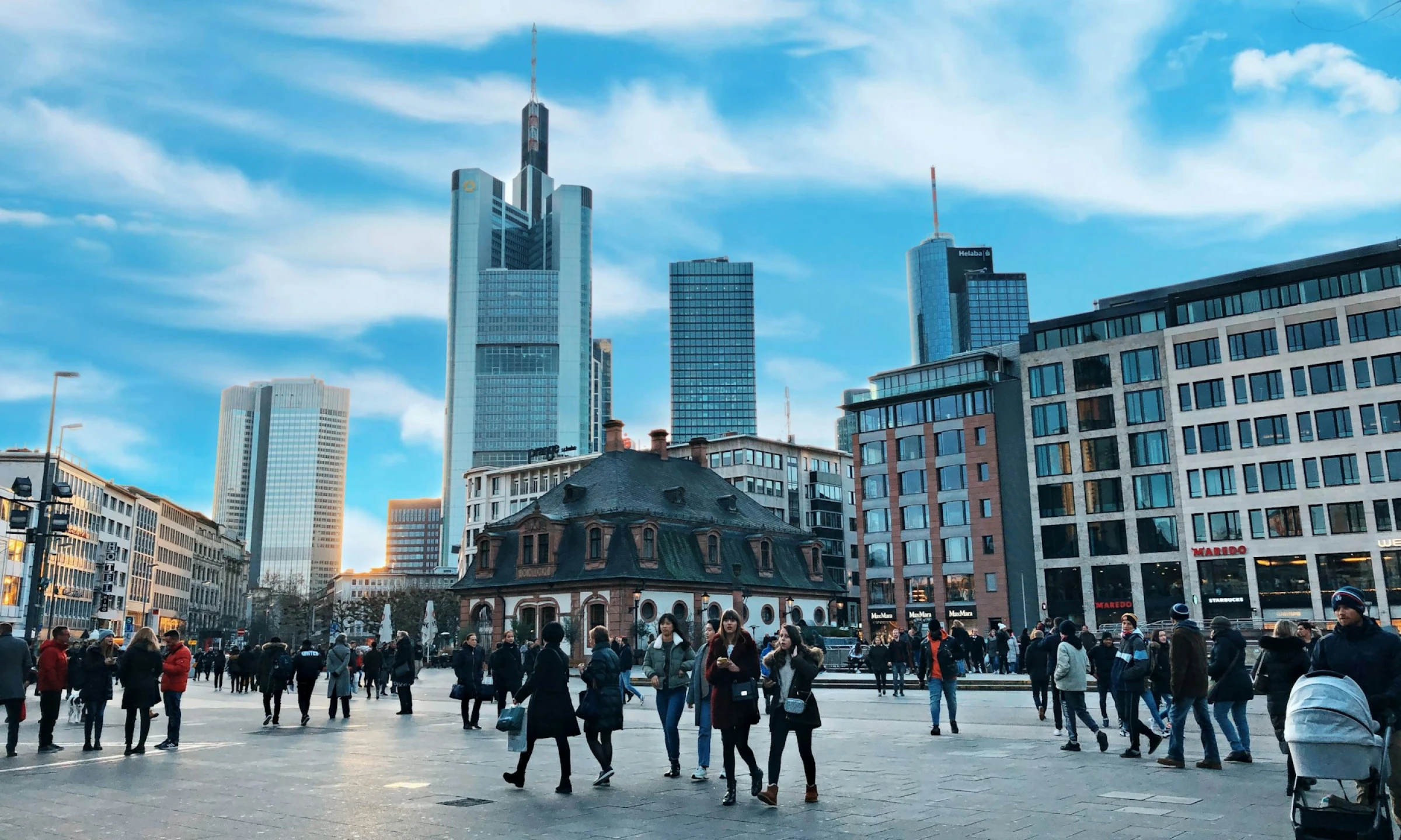 A busy square in Frankfurt with people walking and sightseeing, featuring modern skyscrapers and the historic Hauptwache building under a bright blue winter sky.