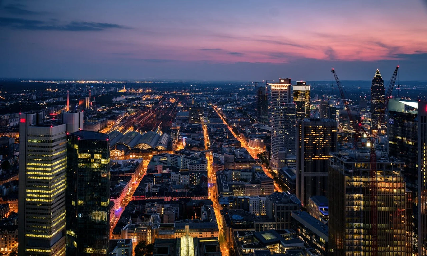Aerial view of Frankfurt at dusk, showcasing illuminated streets, high-rise buildings, and the central train station with a purple and orange sky in the background.