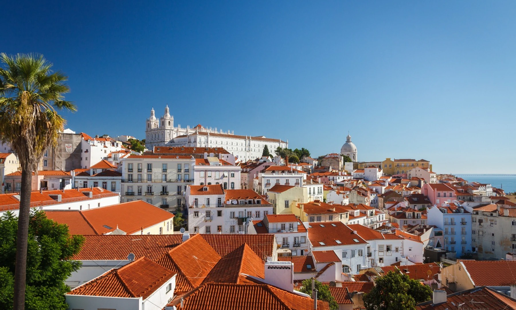 The scenic Alfama district in Lisbon, Portugal, with white buildings and red-tiled roofs under a clear blue sky, featuring the historic São Vicente de Fora monastery in the background.