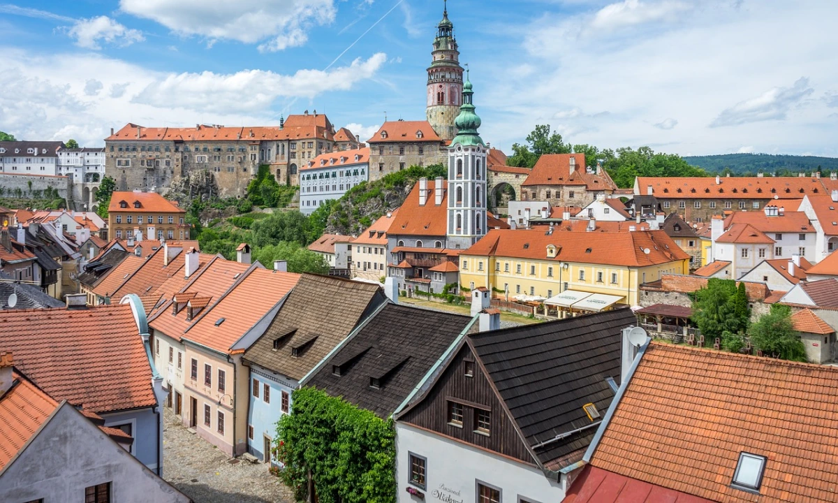 Český Krumlov in the Czech Republic, featuring its historic castle complex, pastel-coloured houses with red-tiled roofs, and a tall tower against a blue sky.